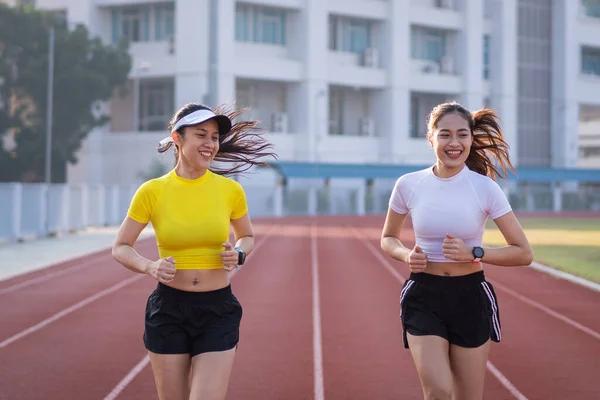 Spor kıyafetleri içinde iki genç Asyalı kadın sağlıklı ve formda kalmak için güneşli bir sabahta stadyumda koşu pistinde koşuyorlar. Genç fitness kadınları stadyum pistinde koşuyor. Spor ve eğlence.