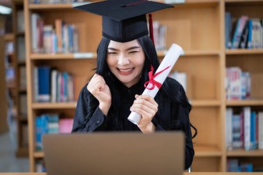 Genç bir Asyalı bayan üniversite mezunu, mezuniyetini kutlamak için sevinç ve heyecan dolu bir şekilde bir dizüstü bilgisayarın önünde ailesine uzaktan bir video konuşması yapıyor.