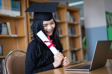 Genç bir Asyalı bayan üniversite mezunu, mezuniyetini kutlamak için sevinç ve heyecan dolu bir şekilde bir dizüstü bilgisayarın önünde ailesine uzaktan bir video konuşması yapıyor.