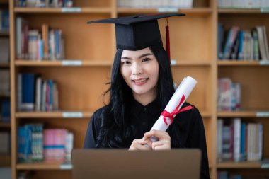 Genç bir Asyalı bayan üniversite mezunu, mezuniyetini kutlamak için sevinç ve heyecan dolu bir şekilde bir dizüstü bilgisayarın önünde ailesine uzaktan bir video konuşması yapıyor.
