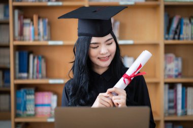 Genç bir Asyalı bayan üniversite mezunu, mezuniyetini kutlamak için sevinç ve heyecan dolu bir şekilde bir dizüstü bilgisayarın önünde ailesine uzaktan bir video konuşması yapıyor.