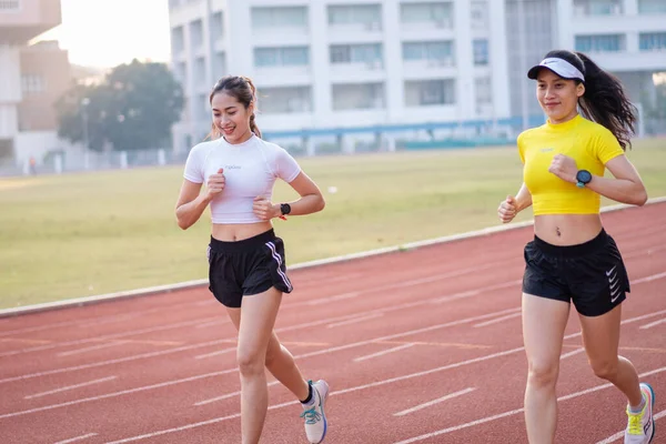 Two young Asian women in sports outfits jogging on running track in city stadium in the sunny morning to keep fitness and healthy lifestyle. Young women run on the stadium track. Sports and recreation