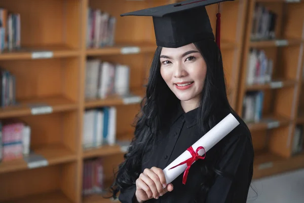 Portrait of a young happy and excited Asian woman university graduates in graduation gown and cap holds and shows a degree certificate to celebrate her education achievement on the commencement day.