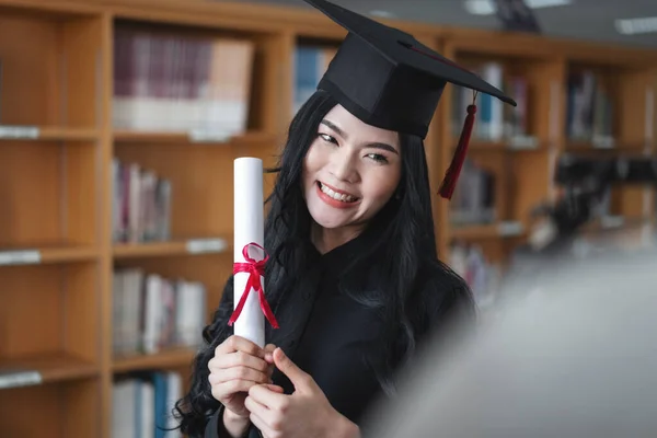 Portrait of a young happy and excited Asian woman university graduates in graduation gown and cap holds and shows a degree certificate to celebrate her education achievement on the commencement day.
