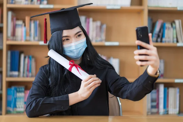 A young cheerful Asian woman university graduates in graduation gown and cap wears a face mask shows a degree certificate via video call to celebrate her education achievement on the commencement day