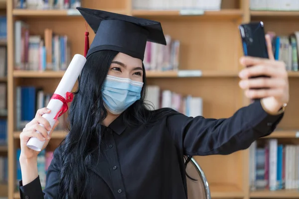 A young cheerful Asian woman university graduates in graduation gown and cap wears a face mask shows a degree certificate via video call to celebrate her education achievement on the commencement day