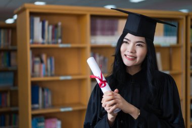 Mezuniyet elbisesi ve maske takan mutlu bir Asyalı kadın üniversitesi mezuniyet törenindeki başarısını kutlamak için diploma tutuyor. Stok fotoğrafı