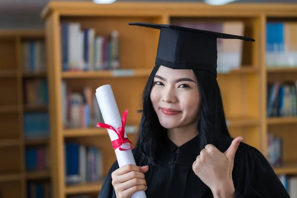 A young happy Asian woman university graduates in graduation gown and cap wears a face mask holds a degree certificate to celebrate her education achievement on the commencement day. Stock photo