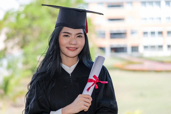 A young happy Asian woman university graduates in graduation gown and cap wears a face mask holds a degree certificate to celebrate her education achievement on the commencement day. Stock photo