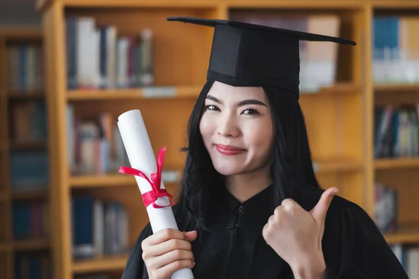 A young happy Asian woman university graduates in graduation gown and cap wears a face mask holds a degree certificate to celebrate her education achievement on the commencement day. Stock photo