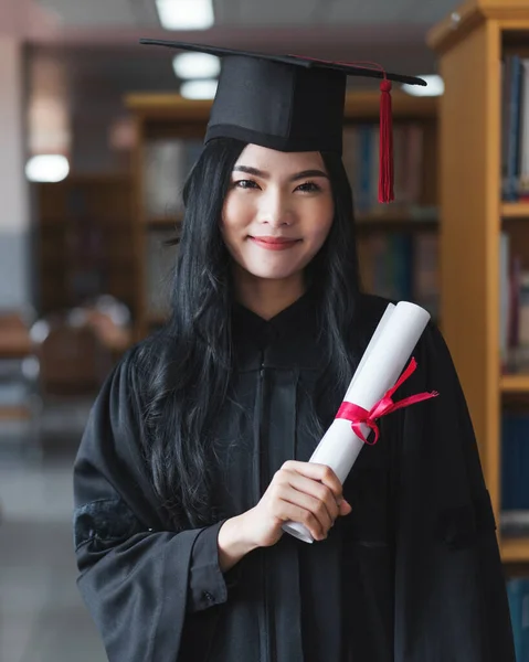 A young happy Asian woman university graduates in graduation gown and cap wears a face mask holds a degree certificate to celebrate her education achievement on the commencement day. Stock photo