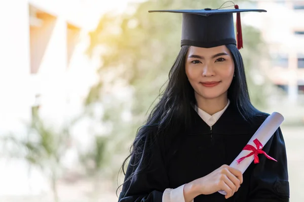 A young happy Asian woman university graduates in graduation gown and cap wears a face mask holds a degree certificate to celebrate her education achievement on the commencement day. Stock photo