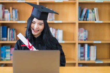 Mezuniyet elbisesi içinde genç bir Asyalı üniversite mezunu. Eğitimini bir dizüstü bilgisayarın önünde kutlamak için sevinç ve heyecan ifade ediyor. Evdeki ailesine uzaktan bir video görüşmesi yapıyor.