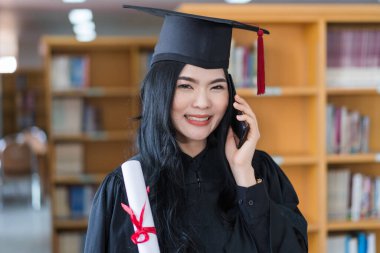 Mezuniyet elbisesi ve şapkası elinde diploması olan neşeli Asyalı genç üniversite mezunu bir kadın mezuniyet törenindeki başarısını kutlamak için arkadaşlarına telefon eder.