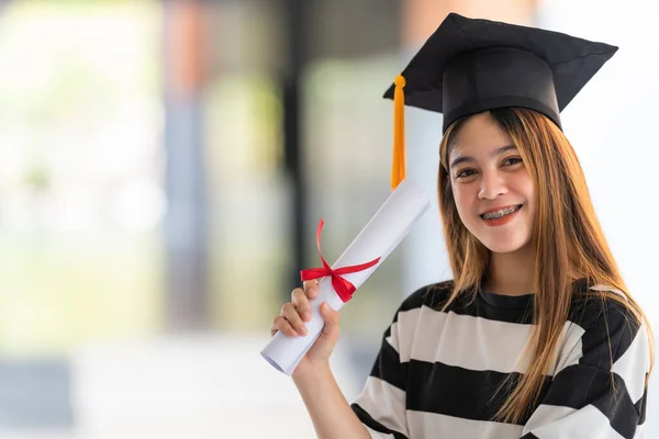 Young asian woman university graduates in graduation gown and mortarboard holds a degree certificate celebrates education achievement in the university campus.  Education stock photo