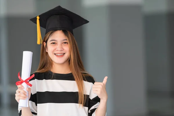 Young asian woman university graduates in graduation gown and mortarboard holds a degree certificate celebrates education achievement in the university campus.  Education stock photo