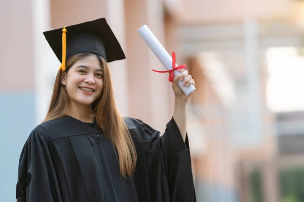 卒業生のガウンとモーターボードを着た若い幸せなアジアの女性大学卒業生は 大学のキャンパスで教育の成果を祝う学位証明書を保持しています 教育ストックフォト — ストック写真