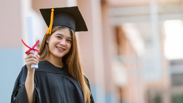 卒業生のガウンとモーターボードを着た若い幸せなアジアの女性大学卒業生は 大学のキャンパスで教育の成果を祝う学位証明書を保持しています 教育ストックフォト — ストック写真