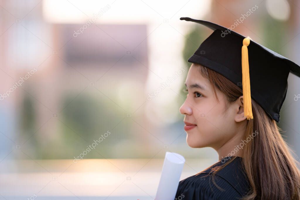 A young happy Asian woman university graduate in graduation gown and mortarboard holds a degree certificate celebrates education achievement in the university campus.  Education stock photo