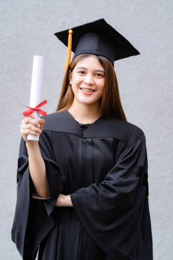Mezuniyet elbisesi ve havan tahtasıyla genç mutlu bir Asyalı kadın üniversite mezunu üniversite kampüsünde eğitim başarısını kutlamak için diploma sahibi. Eğitim stoku fotoğrafı