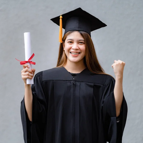 Mezuniyet elbisesi ve havan tahtasıyla genç mutlu bir Asyalı kadın üniversite mezunu üniversite kampüsünde eğitim başarısını kutlamak için diploma sahibi. Eğitim stoku fotoğrafı