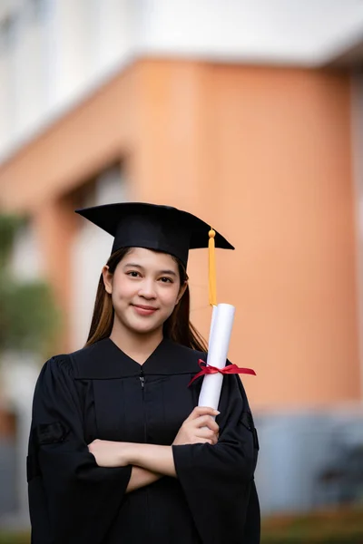 Mezuniyet elbisesi ve havan tahtasıyla genç mutlu bir Asyalı kadın üniversite mezunu üniversite kampüsünde eğitim başarısını kutlamak için diploma sahibi. Eğitim stoku fotoğrafı