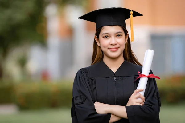 Mezuniyet elbisesi ve havan tahtasıyla genç mutlu bir Asyalı kadın üniversite mezunu üniversite kampüsünde eğitim başarısını kutlamak için diploma sahibi. Eğitim stoku fotoğrafı