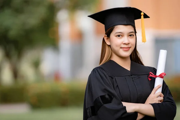 Mezuniyet elbisesi ve havan tahtasıyla genç mutlu bir Asyalı kadın üniversite mezunu üniversite kampüsünde eğitim başarısını kutlamak için diploma sahibi. Eğitim stoku fotoğrafı