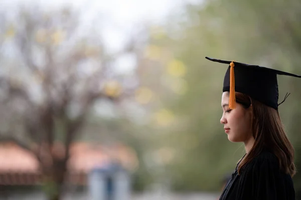 A young happy Asian woman university graduate in graduation gown and mortarboard holds a degree certificate celebrates education achievement in the university campus.  Education stock photo