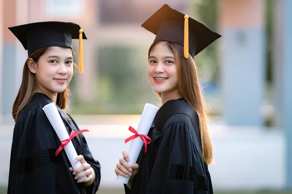 Genç mutlu Asyalı kadın üniversite mezunları mezuniyet elbisesi ve havan tahtasıyla üniversite kampüsünde eğitim başarısını diploma ile kutluyorlar. Eğitim stoku fotoğrafı