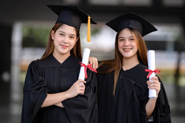 Genç mutlu Asyalı kadın üniversite mezunları mezuniyet elbisesi ve havan tahtasıyla üniversite kampüsünde eğitim başarısını diploma ile kutluyorlar. Eğitim stoku fotoğrafı