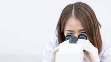 A young scientist woman in a laboratory coat looking through a microscope in a laboratory to do research and experiment. Scientist working in a laboratory. Education stock photo clipart