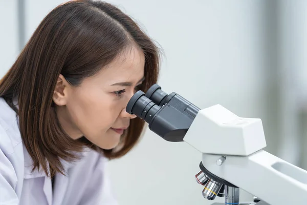 A young scientist woman in a laboratory coat looking through a microscope in a laboratory to do research and experiment. Scientist working in a laboratory. Education stock photo