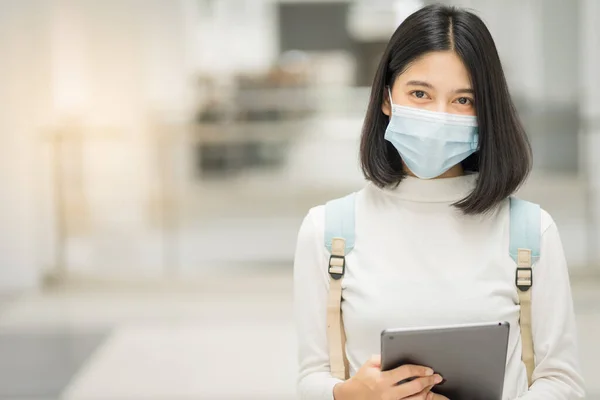 Portrait of a young Asian teenage college student in casual dress, holds tablet with school backpack, wears medical face mask and keeps social distancing during COVID-19 pandemic in college campus