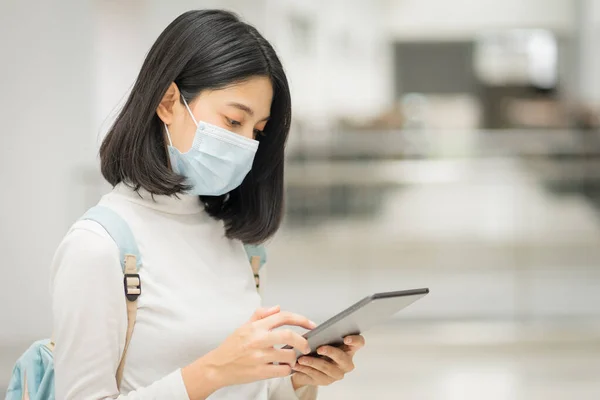 Portrait of a young Asian teenage college student in casual dress, holds tablet with school backpack, wears medical face mask and keeps social distancing during COVID-19 pandemic in college campus