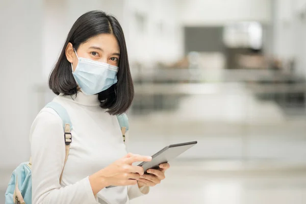 Portrait of a young Asian teenage college student in casual dress, holds tablet with school backpack, wears medical face mask and keeps social distancing during COVID-19 pandemic in college campus