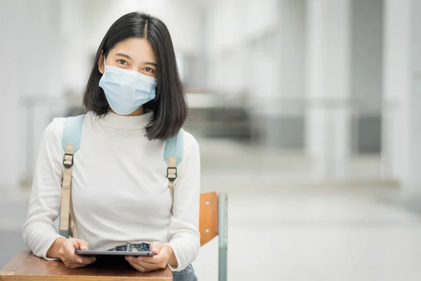 Portrait of a young Asian teenage college student in casual dress, holds tablet with school backpack, wears medical face mask and keeps social distancing during COVID-19 pandemic in college campus