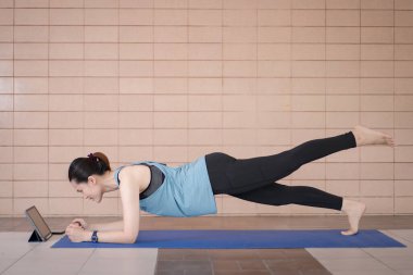Orta yaşlı, rahat spor kıyafetleri içinde bir Asyalı kadın COVID-19 salgını ve şehir tecridi sırasında evdeki bir tablet üzerinde uygulamalı egzersiz programı uyguluyor.
