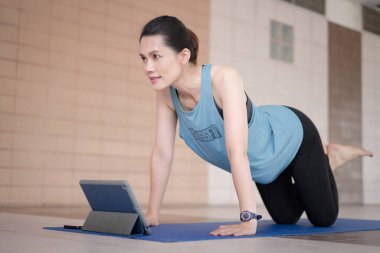 Orta yaşlı, rahat spor kıyafetleri içinde bir Asyalı kadın COVID-19 salgını ve şehir tecridi sırasında evdeki bir tablet üzerinde uygulamalı egzersiz programı uyguluyor.