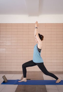 Orta yaşlı, rahat spor kıyafetleri içinde bir Asyalı kadın COVID-19 salgını ve şehir tecridi sırasında evdeki bir tablet üzerinde uygulamalı egzersiz programı uyguluyor.