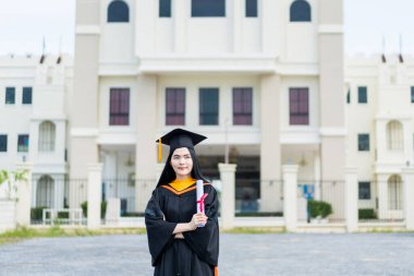 Mezuniyet elbisesi ve havan tahtasıyla güzel bir Asyalı kadın üniversite mezunu üniversite mezuniyetine katıldıktan sonra üniversite binasının önünde diploma tutuyor.