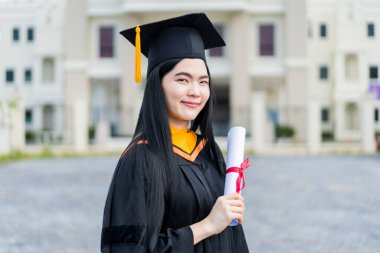 Mezuniyet elbisesi ve havan tahtasıyla güzel bir Asyalı kadın üniversite mezunu üniversite mezuniyetine katıldıktan sonra üniversite binasının önünde diploma tutuyor.