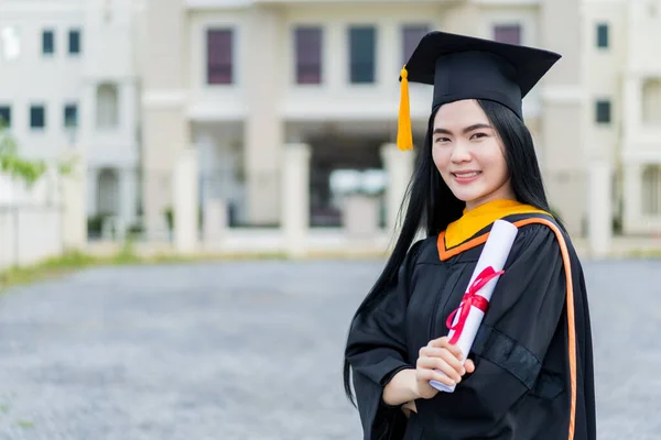 卒業生のガウンとモーターボードを着た若い美しいアジアの女性大学卒業生は 大学の卒業式に参加した後 大学の建物の前に学位証明書スタンドを保持しています — ストック写真