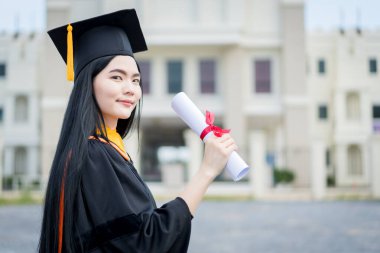 Mezuniyet elbisesi ve havan tahtasıyla güzel bir Asyalı kadın üniversite mezunu üniversite mezuniyetine katıldıktan sonra üniversite binasının önünde diploma tutuyor.