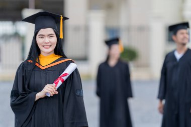 Mezuniyet elbisesi ve havan tahtasıyla güzel bir Asyalı kadın üniversite mezunu üniversite mezuniyetine katıldıktan sonra üniversite binasının önünde diploma tutuyor.