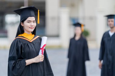 Mezuniyet elbisesi ve havan tahtasıyla güzel bir Asyalı kadın üniversite mezunu üniversite mezuniyetine katıldıktan sonra üniversite binasının önünde diploma tutuyor.
