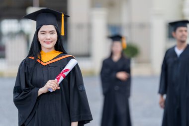 Mezuniyet elbisesi ve havan tahtasıyla güzel bir Asyalı kadın üniversite mezunu üniversite mezuniyetine katıldıktan sonra üniversite binasının önünde diploma tutuyor.