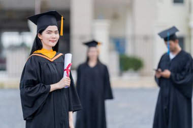 Mezuniyet elbisesi ve havan tahtasıyla güzel bir Asyalı kadın üniversite mezunu üniversite mezuniyetine katıldıktan sonra üniversite binasının önünde diploma tutuyor.