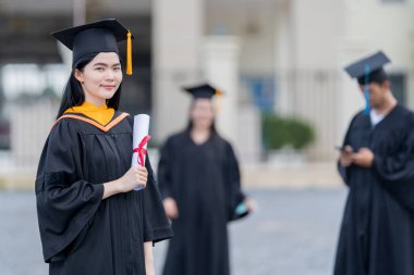 Mezuniyet elbisesi ve havan tahtasıyla güzel bir Asyalı kadın üniversite mezunu üniversite mezuniyetine katıldıktan sonra üniversite binasının önünde diploma tutuyor.
