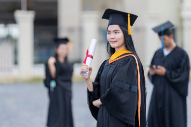 Mezuniyet elbisesi ve havan tahtasıyla güzel bir Asyalı kadın üniversite mezunu üniversite mezuniyetine katıldıktan sonra üniversite binasının önünde diploma tutuyor.